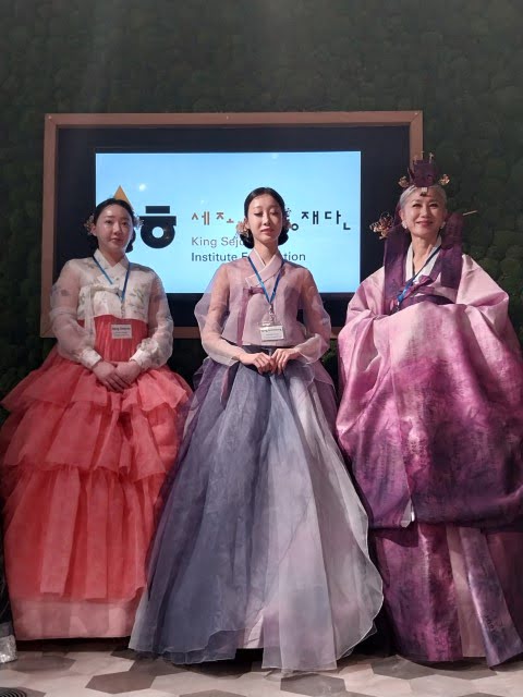 Three people in traditional Korean attire, known as hanbok, stand before a screen displaying text related to the King Sejong Institute Foundation.