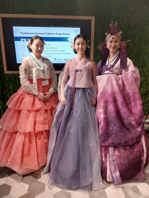 Three people in traditional Korean attire stand before an electronic display promoting cultural experiences. They are wearing colorful hanboks with intricate designs.